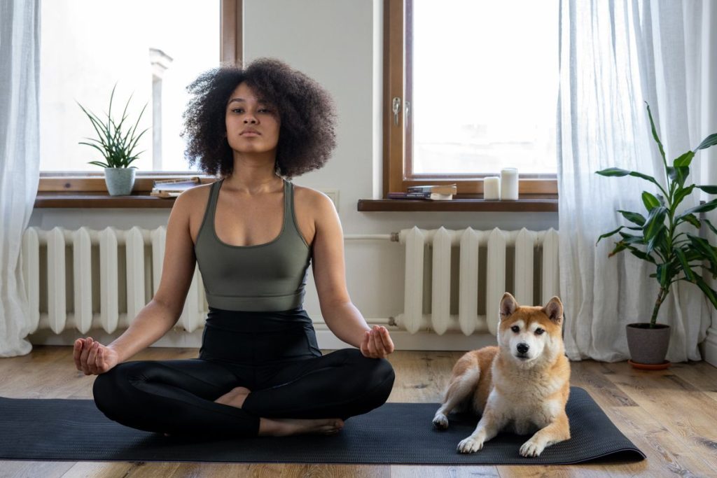 Femme qui médite sur son tapis dans son salon, son chien installé à côté d'elle